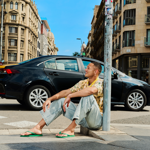 Homem usando chinelos verdes em calçada urbana, com carro e prédios ao fundo